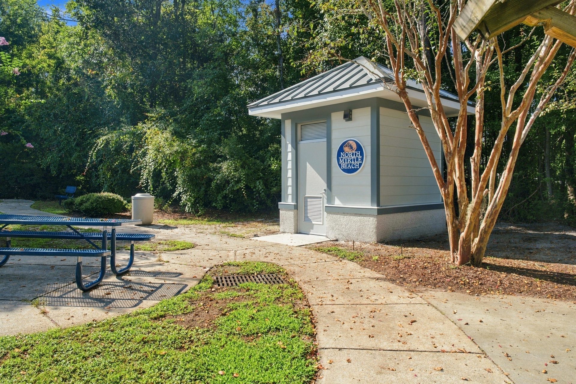 Public restroom building in a North Myrtle Beach city park, with the concrete walkway curving past it and picnic tables under the trees
